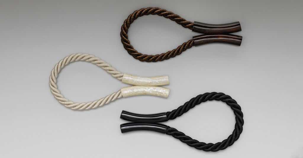 Ein sehr außergewöhnlicher Raffhalter mit Magnetverschluss. Die beiden Endstücke sind in den Kolorits Cremeweiß und Schokolade aus Perlmutt-Intarsien gefertigt; in Schwarz sind die Endstücke hochglänzend lackiert. Die gedrehte Kordel aus Acetatfäden zeigt einen feinen Striée-Effekt.